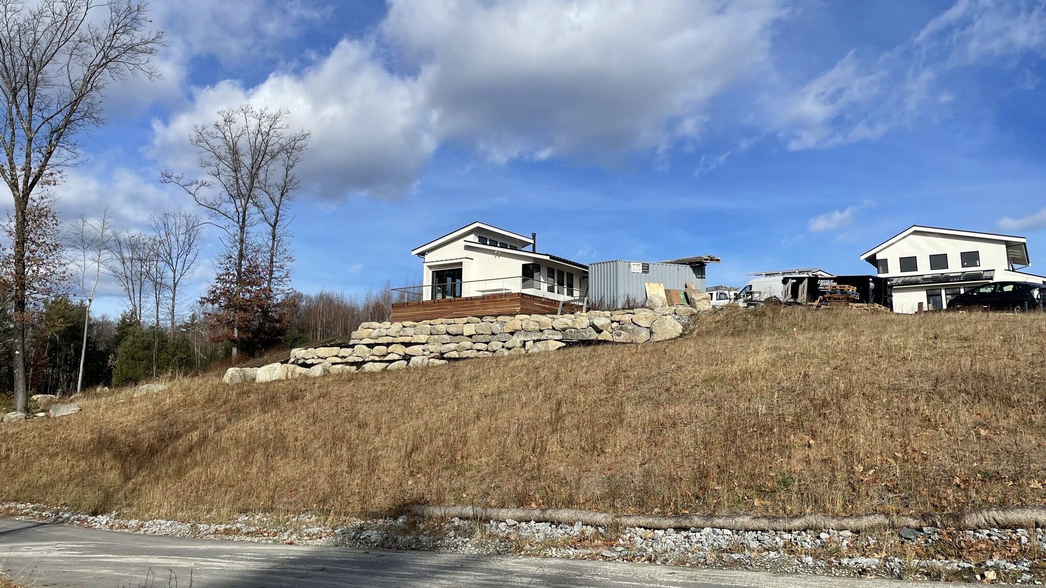 Modern home under construction sit atop a sloped, grassy hillside, with a large stacked-stone retaining wall and wooden deck in the foreground and a coir erosion-control log installed along the base of the slope beside a gravel road.